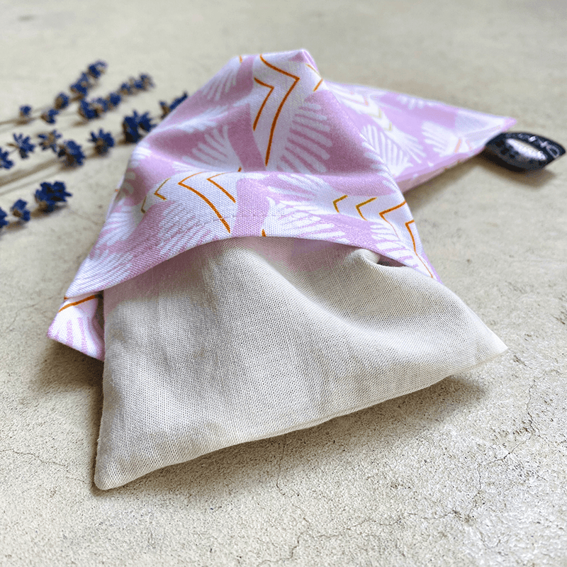 Coussin relaxant pour les yeux de la marque Belle Luce, au motif violet et orange avec des motifs géométriques. Ce coussin est utilisé pour la relaxation, l'aromathérapie et la méditation, offrant des options réfrigérantes ou chauffantes. L'emballage met en avant ses bienfaits de détente. Des brins de lavande sont posés à côté, soulignant le lien avec l'aromathérapie